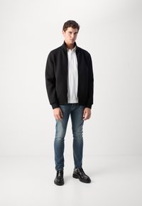 Fekete, zippzáros dzseki magas gallérral, fehér póló fölött. Világoskék, slim-fit farmert és fekete bokacsizmát visel.
