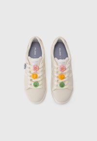 Beige sneakers with a smooth texture, featuring decorative charms: a pink flower, orange heart, and green smiley face on the laces.