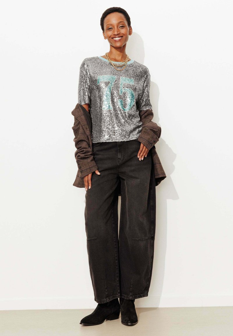 Chemise à manches courtes argentée ornée de sequins avec le numéro « 75 » en turquoise, associée à un pantalon noir fluide taille haute et des bottines noires.
