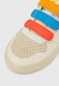 Biały skórzany tenisówka z beżowym zamszowym noskiem, wyposażona w kolorowe rzepy w kolorze pomarańczowym, niebieskim i żółtym. Na nosku znajdują się perforowane detale.