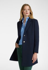Blazer azul marino con un diseño ajustado, botones dorados y un bolsillo. Llevado sobre una camisa de rayas azul claro y pantalones verdes.