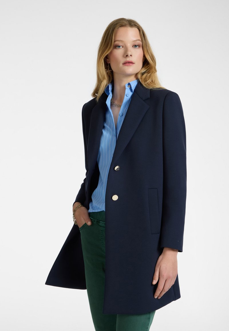 Blazer azul marino con un diseño ajustado, botones dorados y un bolsillo. Llevado sobre una camisa de rayas azul claro y pantalones verdes.