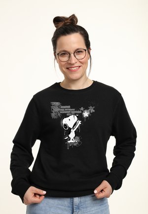 Donna sorridente con occhiali indossa una felpa nera con Snoopy che raggiunge una stella su uno sfondo in stile graffiti.