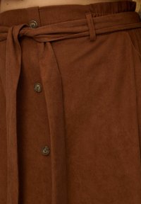 Bruine corduroy rok met een gestrikt riem, voorzien van knopen aan de voorkant en geplooide details langs de zijkanten. Textiel met een zachte afwerking.