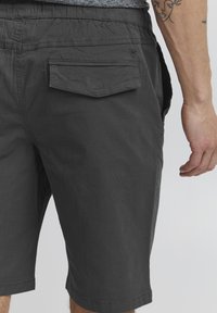 Shorts gris charbon en tissu léger, dotés d'une poche arrière avec un rabat et un détail de marque subtil. Texture lisse et longueur jusqu'au genou.