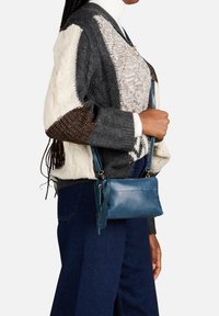 Blauwe leren schoudertas, rechthoekige vorm, ritssluiting, verstelbare schouderband, met een decoratieve label. Gedragen met een gebreide trui met meerdere patronen.