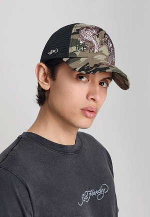 Ung mand iført en sort mesh camouflage kasket med dragebroderi og en mørkegrå Ed Hardy skjorte, der vender mod venstre på en grå baggrund.
