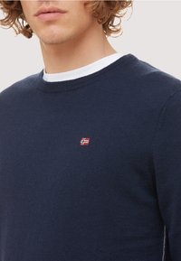 Pull en maille bleu marine avec un col rond, présentant un petit emblème de drapeau rouge et blanc sur la poitrine et un tissu texturé.