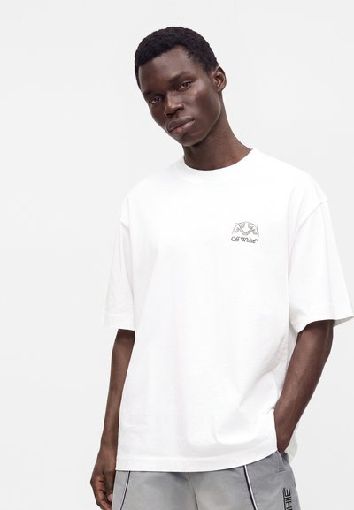 Άνδρας που φοράει λευκό t-shirt Off-White και ανοιχτό γκρι παντελόνι, στέκεται με το ένα χέρι στην τσέπη του μπροστά σε μονόχρωμο φόντο.