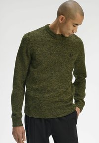 Maglione verde lavorato a maglia con finitura testurizzata, scollo rotondo e polsini a coste. Indossato con pantaloni neri, che mostrano uno stile rilassato.