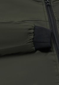 Grüner gepolsterter Jackenstoff mit glatter Textur, mit einem schwarzen elastischen Bündchen und einem Reißverschluss an der Vorderseite.