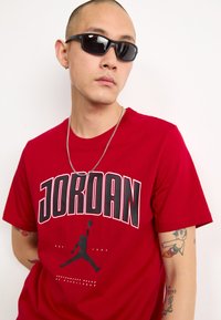 Crvena pamučna majica s podebljanim natpisom "JORDAN" i grafičkim siluetom košarkaša. Dodatna oprema uključuje srebrnu ogrlicu i crne sunčane naočale.