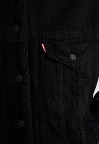 Czarna kurtka dżinsowa z zatrzaskami, kieszenią na klatce piersiowej i czerwoną metką logo. Posiada teksturowany materiał i zbudowaną konstrukcję.