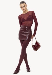 Bordeaux doorzichtig langemouwtop met versieringen, gecombineerd met een glanzende leren minirok en bijpassende panty; houdt een kleine handtas vast.