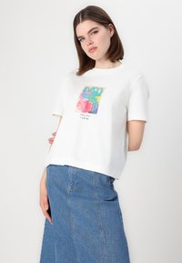 T-shirt blanc à manches courtes avec un imprimé abstrait coloré et du texte. Associé à une jupe en denim, présentant des coutures latérales visibles et des poches.