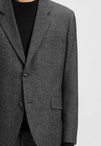 Gray wool blazer, single-breasted design, two buttons, notch lapels, textured fabric, chest pocket, worn over a black top.