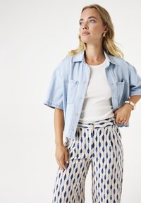 Camisa de mezclilla de manga corta en azul claro sobre una camiseta ajustada blanca, combinada con pantalones crema con un patrón de hojas azules.