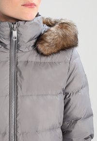 Giacca puffer grigia con colletto alto e rifinitura in pelliccia sintetica. Cerniera argentata e texture trapuntata con dettagli di cucitura orizzontale.