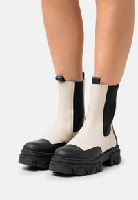 Kreminės odinės masyvios kulkšnės botai su juodais akcentais. Ypatybės: elastiniai šonai, traukimo kilpos ir tekstūruoti guminiai padai.