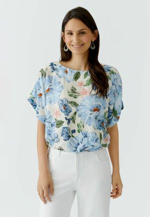 Frau lächelt, trägt eine weiße Bluse mit blauen und rosa Blumenmustern und weiße hoch sitzende Hosen, vor einem schlichten hellen Hintergrund.