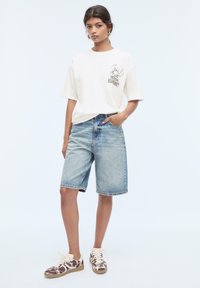 T-shirt en coton blanc avec impression graphique sur la poitrine, associé à un short en denim bleu clair. Baskets brunes à motifs avec lacets crème.