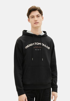 Sudadera negra con un bolsillo frontal, presenta el texto "DENIM TOM TAILOR" en blanco y rosa dorado junto con la fecha 2007. Tela suave con capucha ajustable.