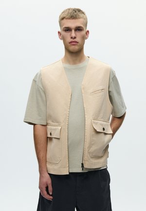 Giovane uomo con capelli biondi corti che indossa un gilet beige sopra una maglietta grigio chiaro e pantaloni neri, in piedi contro uno sfondo chiaro e uniforme.