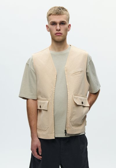 Młody mężczyzna z krótkimi blond włosami, ubrany w beżową kamizelkę roboczą na jasnoszarej koszulce i czarnych spodniach, stojący na tle jasnego, jednolitego tła.