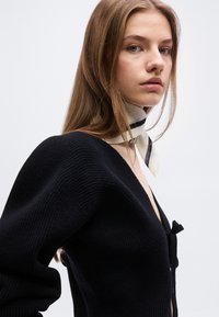 Νέα γυναίκα με μακριά ανοιχτό καστανά μαλλιά, φορώντας μαύρο πουλόβερ με βαθύ V λαιμόκοψη και λευκό κασκόλ με μαύρες ρίγες γύρω από το λαιμό της.