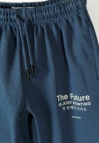 Pantalon avec taille élastique bleue, cordon de serrage et texte blanc indiquant « The Future is Just Starting » avec des caractères coréens en dessous sur la cuisse.