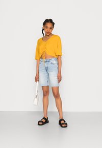 Top amarelo cropped com decote em V e detalhe com cordão, combinado com calções de ganga azul claro e sandálias pretas, segurando uma pequena bolsa branca.