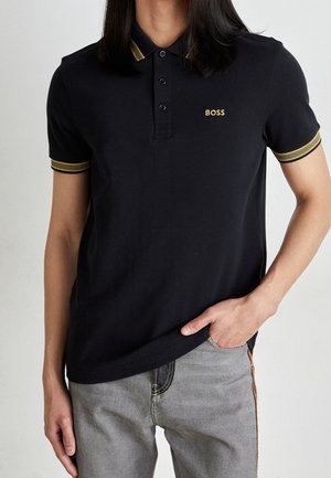 Persoon die een zwart poloshirt draagt met gouden bies op de kraag en mouwen, "BOSS" logo op de borst, en lichtgrijze spijkerbroek met hand in de zak.