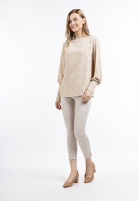 Blouse in zacht beige met een gestructureerd patroon, ballonmouwen en een ronde halslijn. Gecombineerd met strakke lichtbeige broeken en blokhakken.