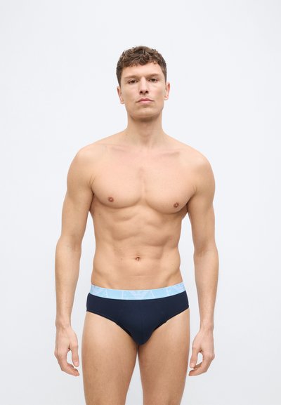 Κατάλληλος νεαρός άνδρας που στέκεται, φοράει σκουρόχρωμο μπλε εσώρουχο με ανοιχτό μπλε ζώνη μέσης, μπροστά σε απλό λευκό φόντο.