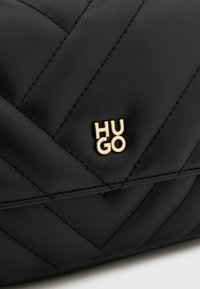 Čierna prešívaná kožená kabelka s logom "HUGO" v zlatej farbe. Diagonálny stehový vzor pridáva dizajnu textúru a hĺbku.