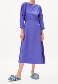 Knee-length, jasně fialové šaty s dlouhými rukávy a kulatým výstřihem, doplněné modrými slip-on sandály s uzlovým designem.