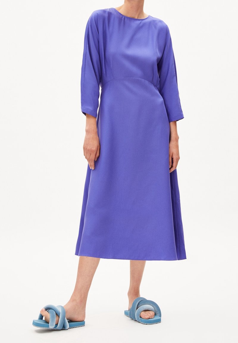 Knee-length, jasně fialové šaty s dlouhými rukávy a kulatým výstřihem, doplněné modrými slip-on sandály s uzlovým designem.