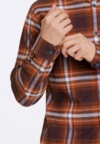 Camicia a quadri con bottoni in tonalità marrone, arancione e bianco, dal tessuto morbido. I polsini sono arrotolati, mostrando bottoni blu.