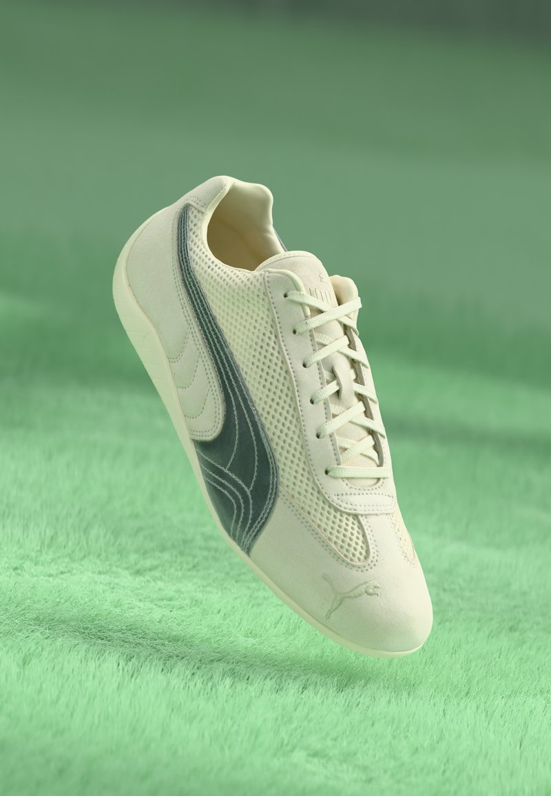 Lys beige Puma sneakers med mørkegrå side stribe, mesh-paneler og snørebånd, svævende over en grøn struktureret overflade.