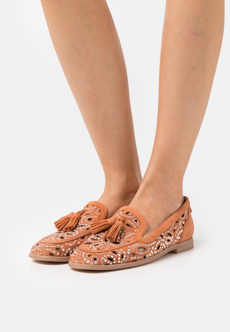 Mocassins en suède beige avec des découpes, ornés de perles blanches et de glands à l'avant, semelle plate et bout arrondi.