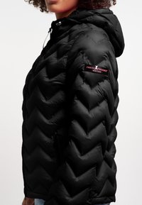 Piumino nero con motivo a zigzag, cappuccio e tasche laterali con cerniera. L'etichetta del marchio è visibile sulla spalla sinistra.