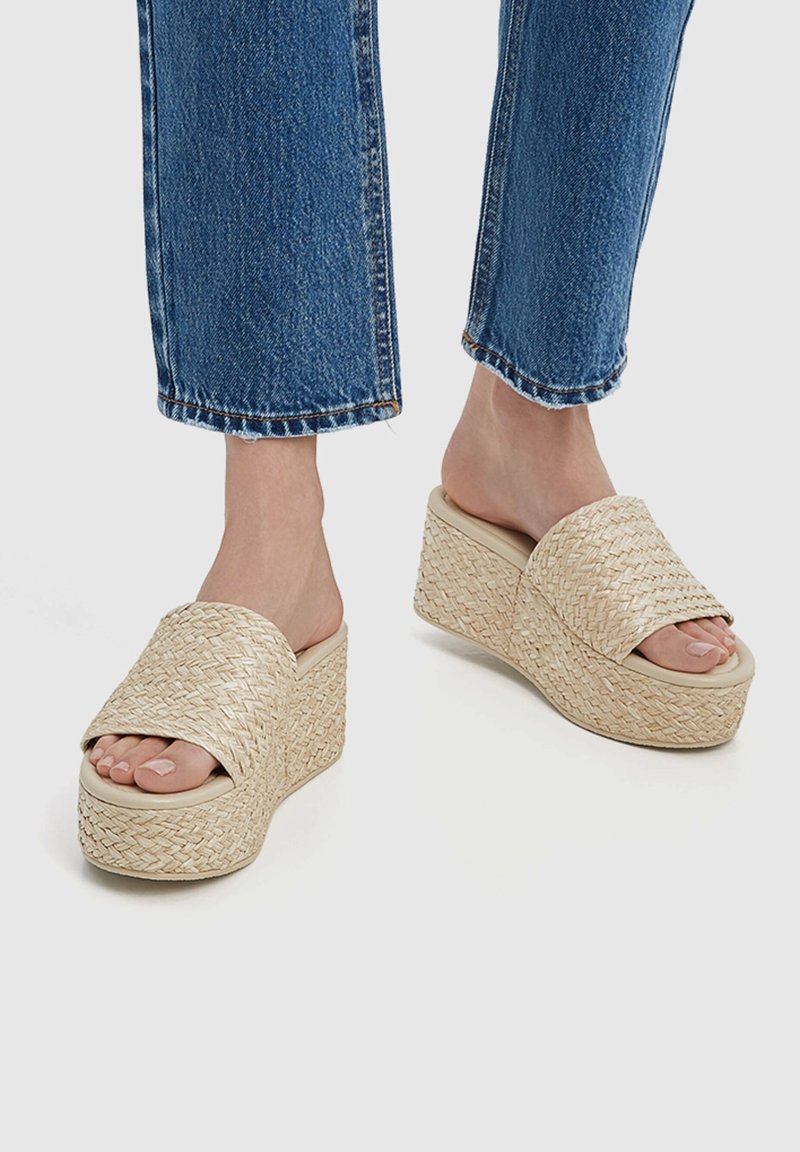 Pieds portant des sandales plates à plateforme tissées beige associées à un jean en denim bleu, présentés sur un fond clair uni.
