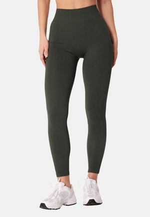 Kobieta ubrana w ciemnozielone legginsy z wysokim stanem i białe buty sportowe, stoi z rękami luźno opuszczonymi wzdłuż ciała na jednolitym tle.