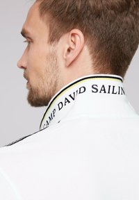 Fehér polo póló fekete és sárga csíkkal ellátott gallérral, rajta hímzett "CAMP DAVID SAILING" felirattal. Közeli nézet hátulról.