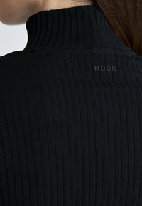 Gros plan d'une personne portant un pull à col roulé côtelé noir avec le mot "HUGO" imprimé près du col au dos.