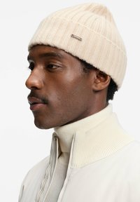 Cremefarvet ribbet strikbeanie med en lille metallisk accent, båret af en person i en lyse farvet zip-up jakke og kravesweater.