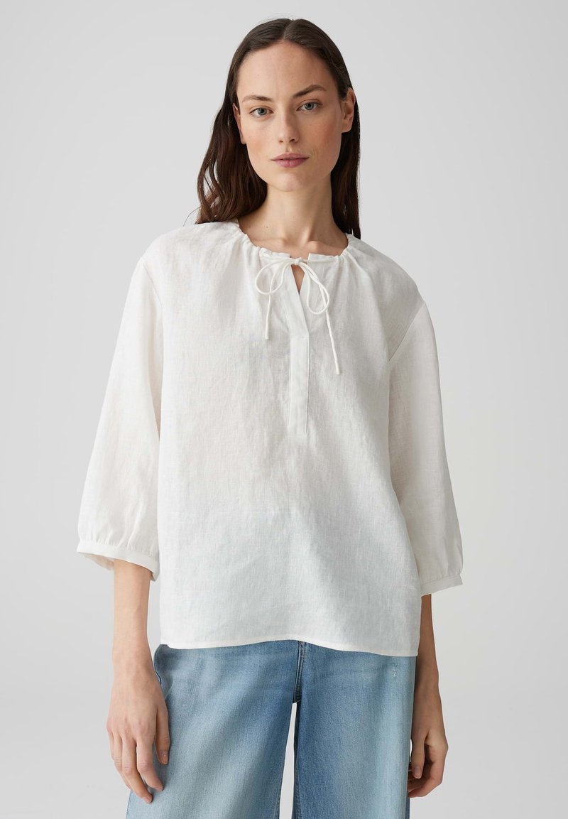 Blusa bianca in lino con scollatura tonda e dettaglio legato. Presenta maniche a tre quarti e una vestibilità ampia, abbinata a jeans azzurri chiari.