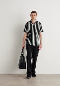Chemise à rayures noires et blanches à manches courtes, boutonnée, avec une poche poitrine, associée à un pantalon noir ample et un sac fourre-tout noir.