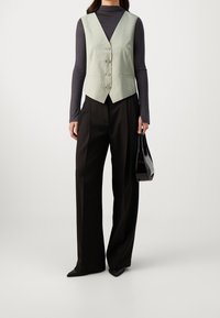 Hellgrüne Weste mit V-Ausschnitt und fünf Knöpfen, kombiniert mit weiten schwarzen Hosen. Als Accessoire wird eine kleine schwarze Handtasche getragen.