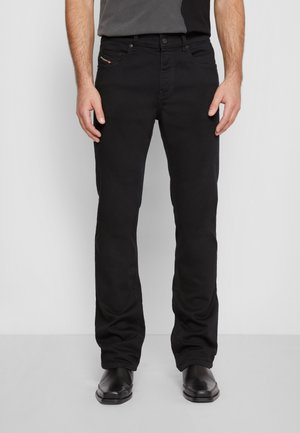 Mann trägt schwarze Jeans mit geradem Bein und schwarze Schuhe, steht vor einem einfarbigen hellgrauen Hintergrund.